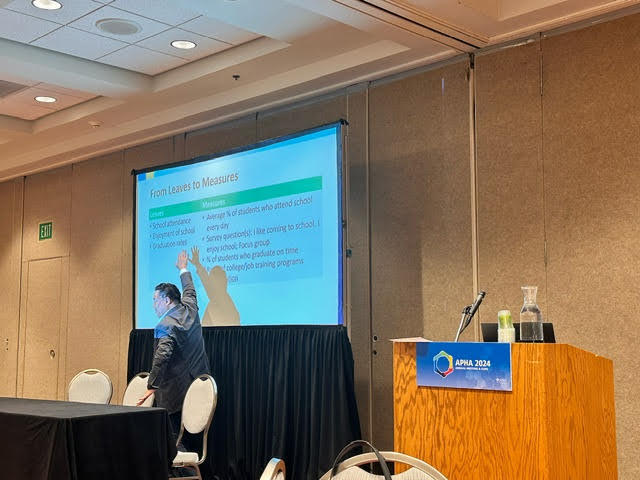 A speaker points to a PowerPoint slide.