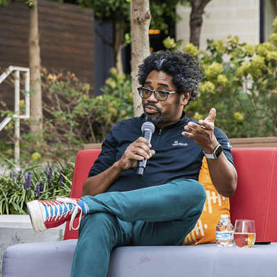 a person with a dark blue shirt and teal pants sits holding a microphone on an outdoor couch 