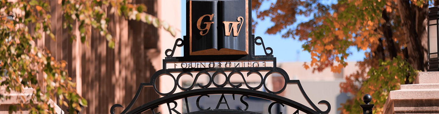 a dark blue GW on a gate surrounded by brown fall leaves on campus
